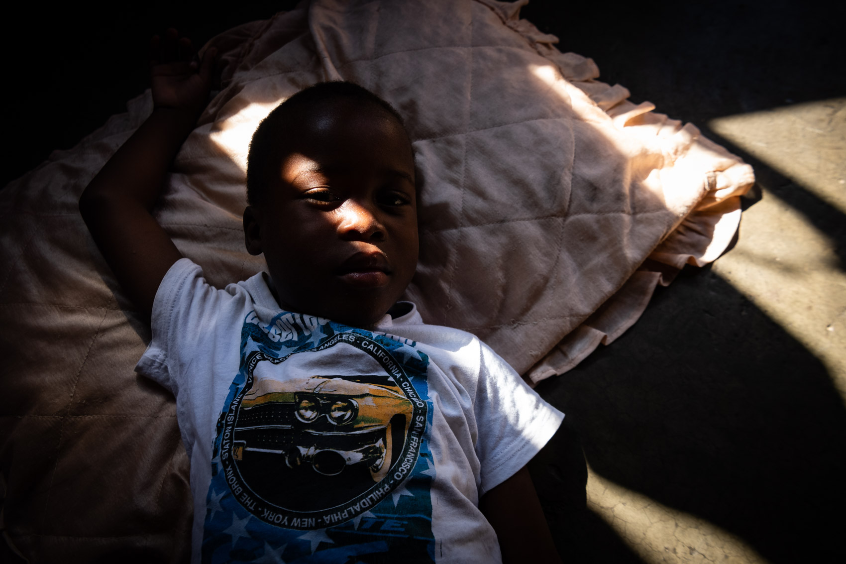 An African boy laying on the ground with a pillow with a ray of light across boy’s face